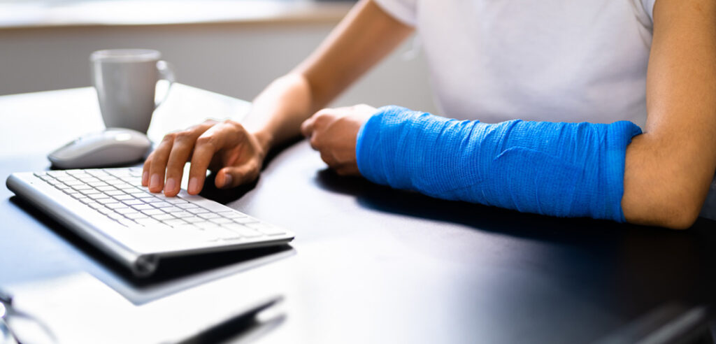 Broken arm in blue cast on worker at computer keyboard. Middle Creek Insurance offers Workers Compensation Insurance in North Carolina